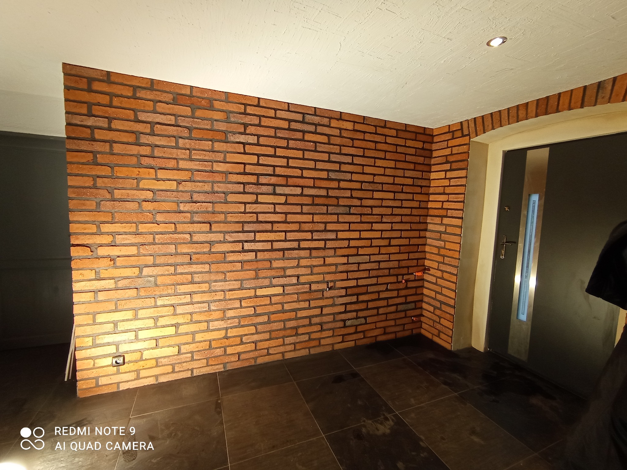 Ściana z cegły klinkierowej wewnątrz pomieszczenia, widoczne fugi i oświetlenie punktowe. Ciemna podłoga z płytek. Fragment drzwi wejściowych.