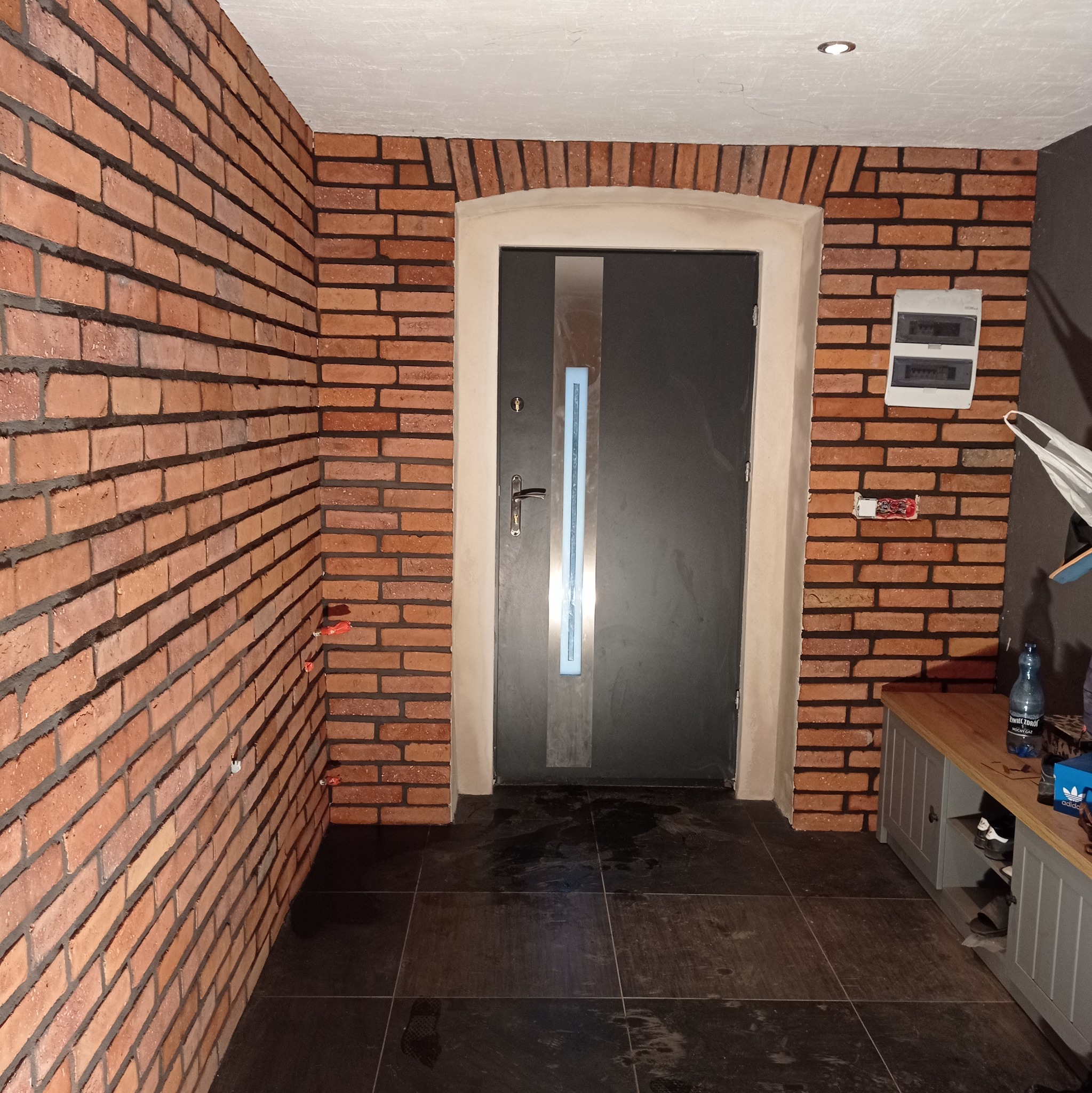 Wykończenie przedpokoju z ceglaną ścianą, szarą stolarką drzwiową i ciemną podłogą. Widoczna szafka na buty i instalacja elektryczna na ścianie.