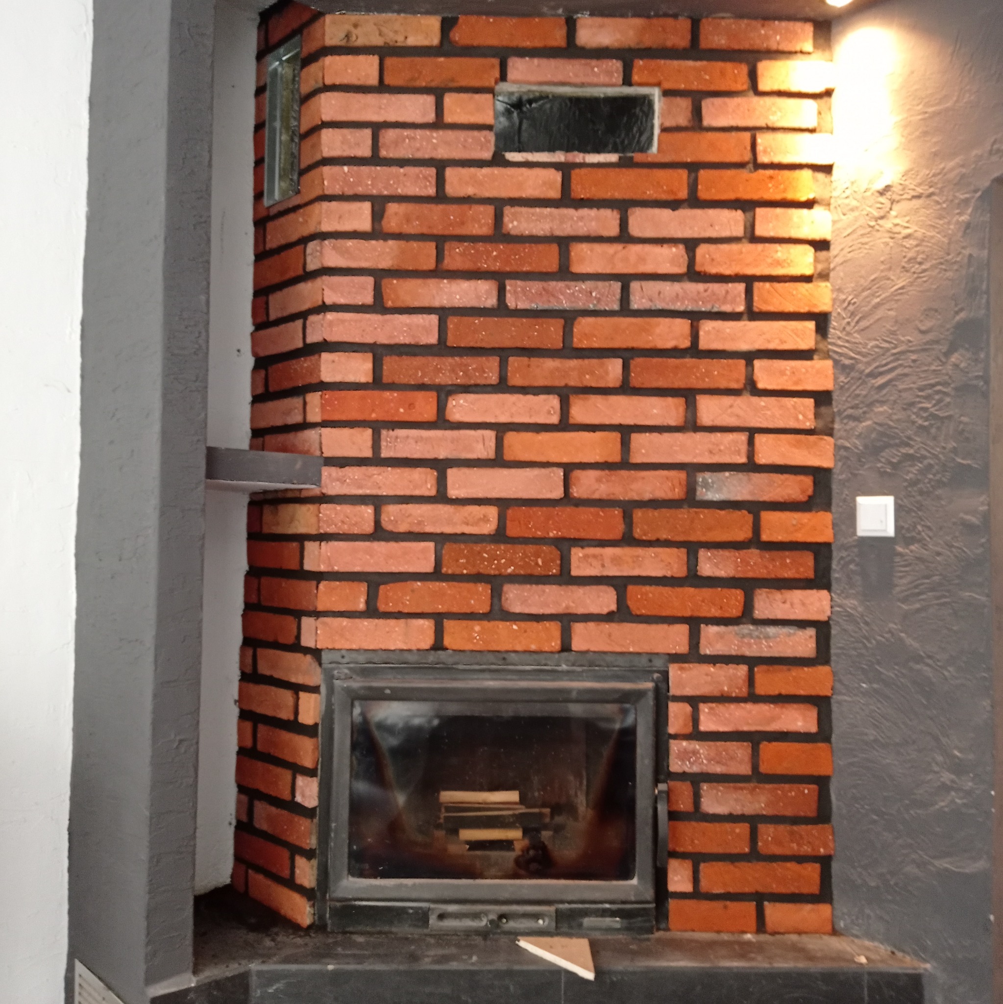 Murowany kominek z czerwonej cegły z czarną fugą, z metalowymi drzwiczkami. Obok ściana z szarą strukturą i włącznik światła. Widoczne drewno wewnątrz kominka.