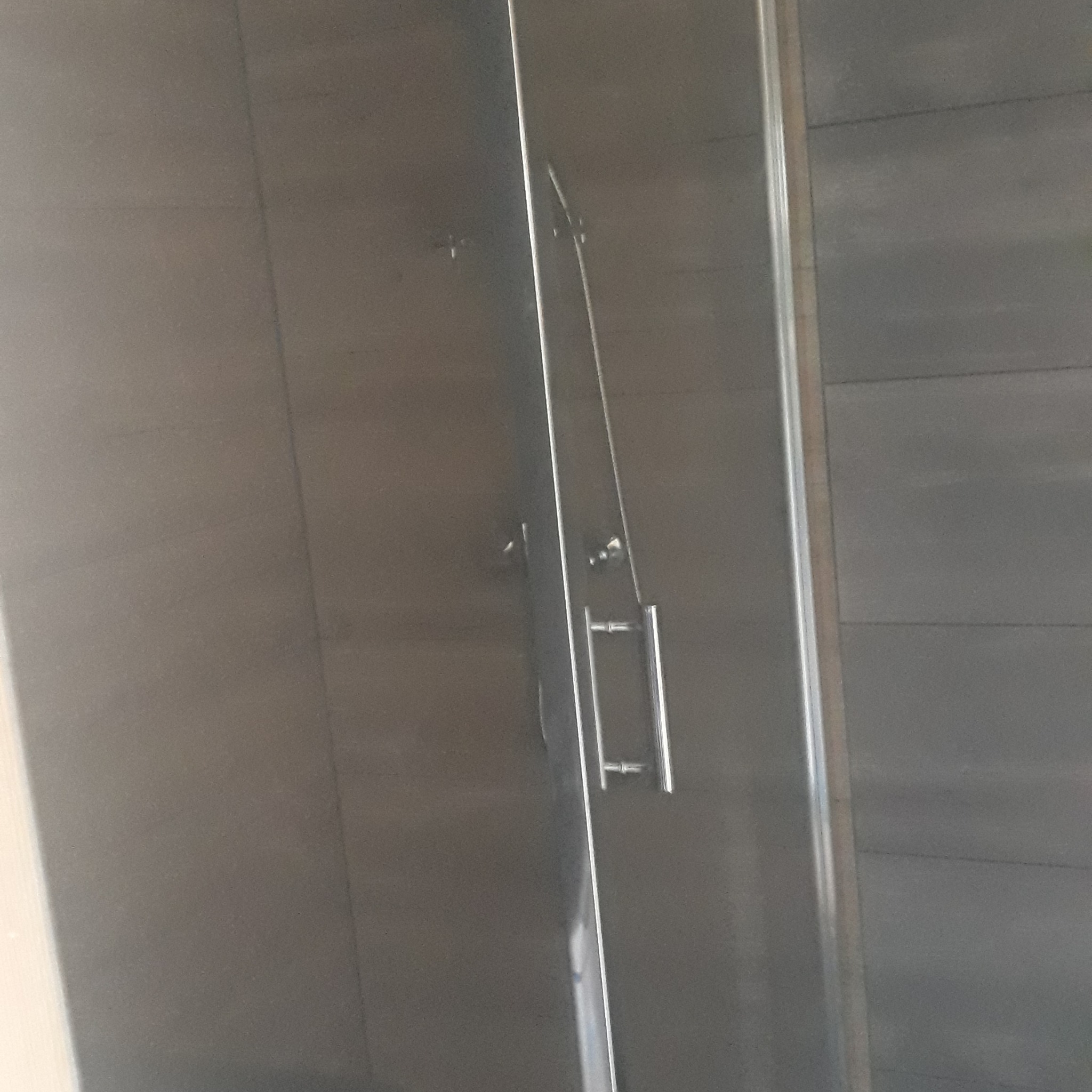 Szklana kabina prysznicowa z chromowanym uchwytem na tle szarych płytek. Minimalistyczny design, widoczne detale mocowań.