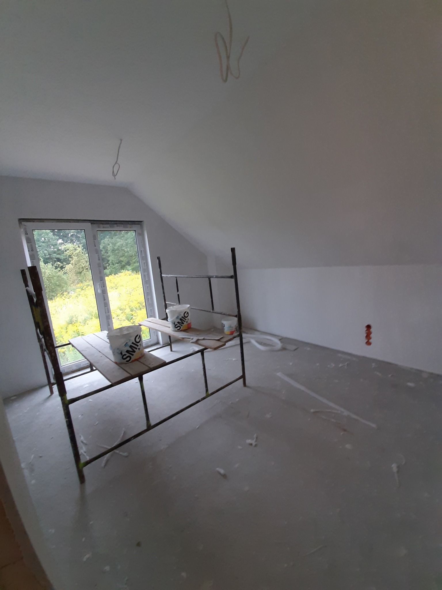 Poddasze w trakcie przygotowania do malowania: widoczne rusztowanie z wiadrami farby SMIG, świeżo zagruntowane ściany i sufit, wystające przewody elektryczne i widok z okna na zieleń.