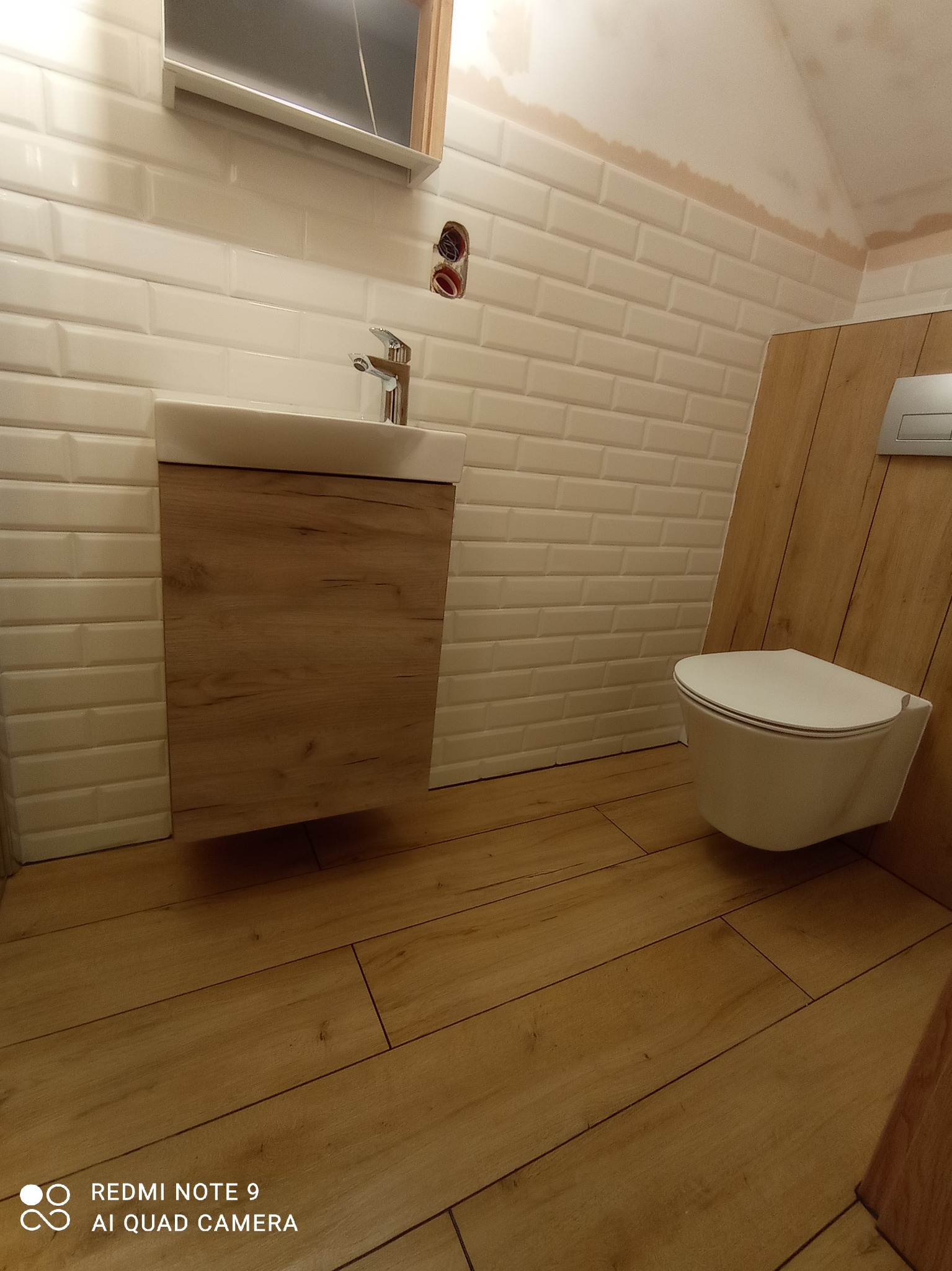 Wykończona łazienka w stylu skandynawskim: białe, cegiełkowate płytki na ścianach, wisząca toaleta z drewnianą obudową, podwieszana szafka z umywalką i drewnopodobną okleiną, oraz podłoga z paneli...