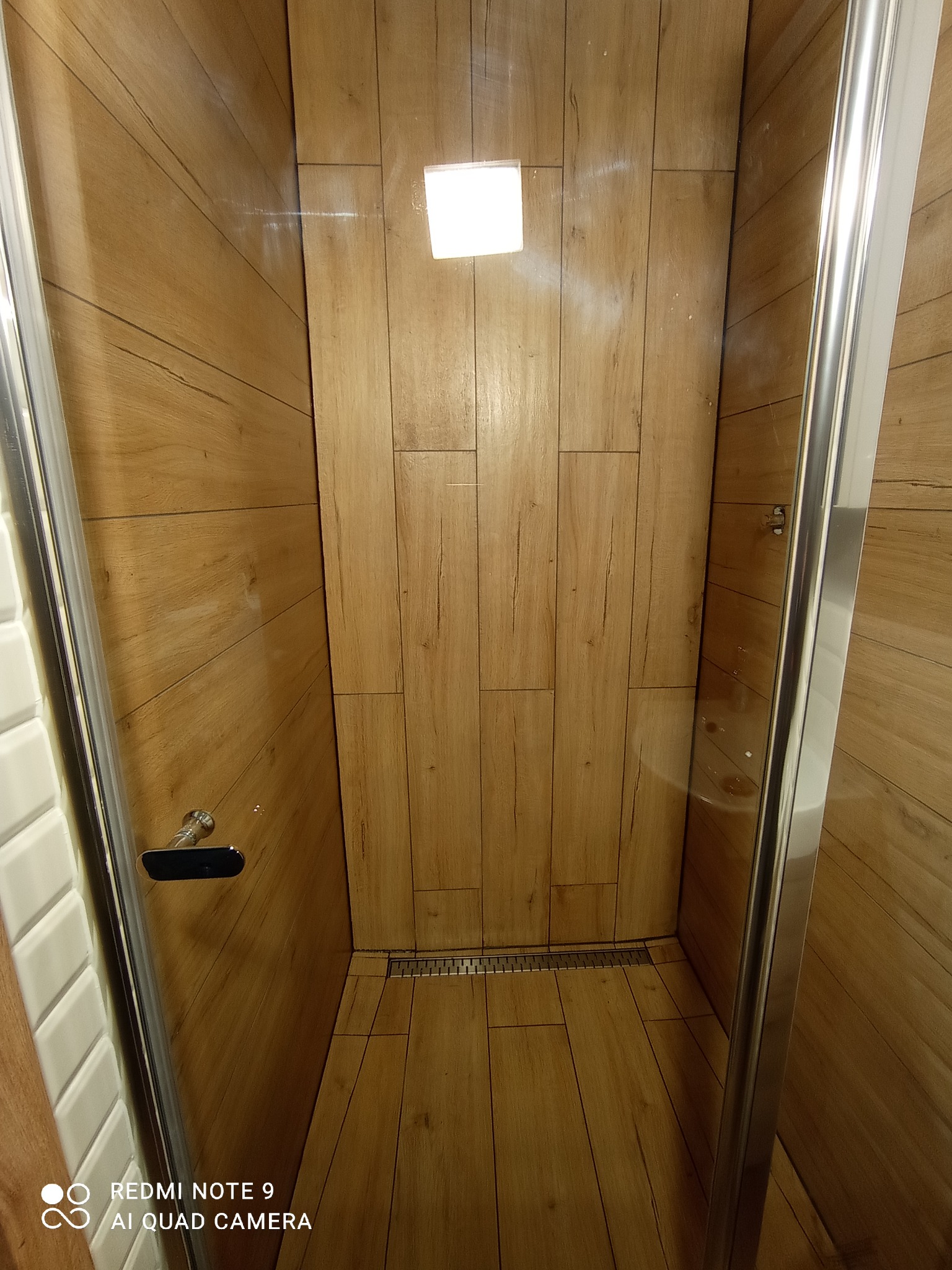 Kabina prysznicowa z wykończeniem imitującym drewno, widoczny odpływ liniowy i chromowane okucia, uchwyt na szklanej ściance po lewej stronie.