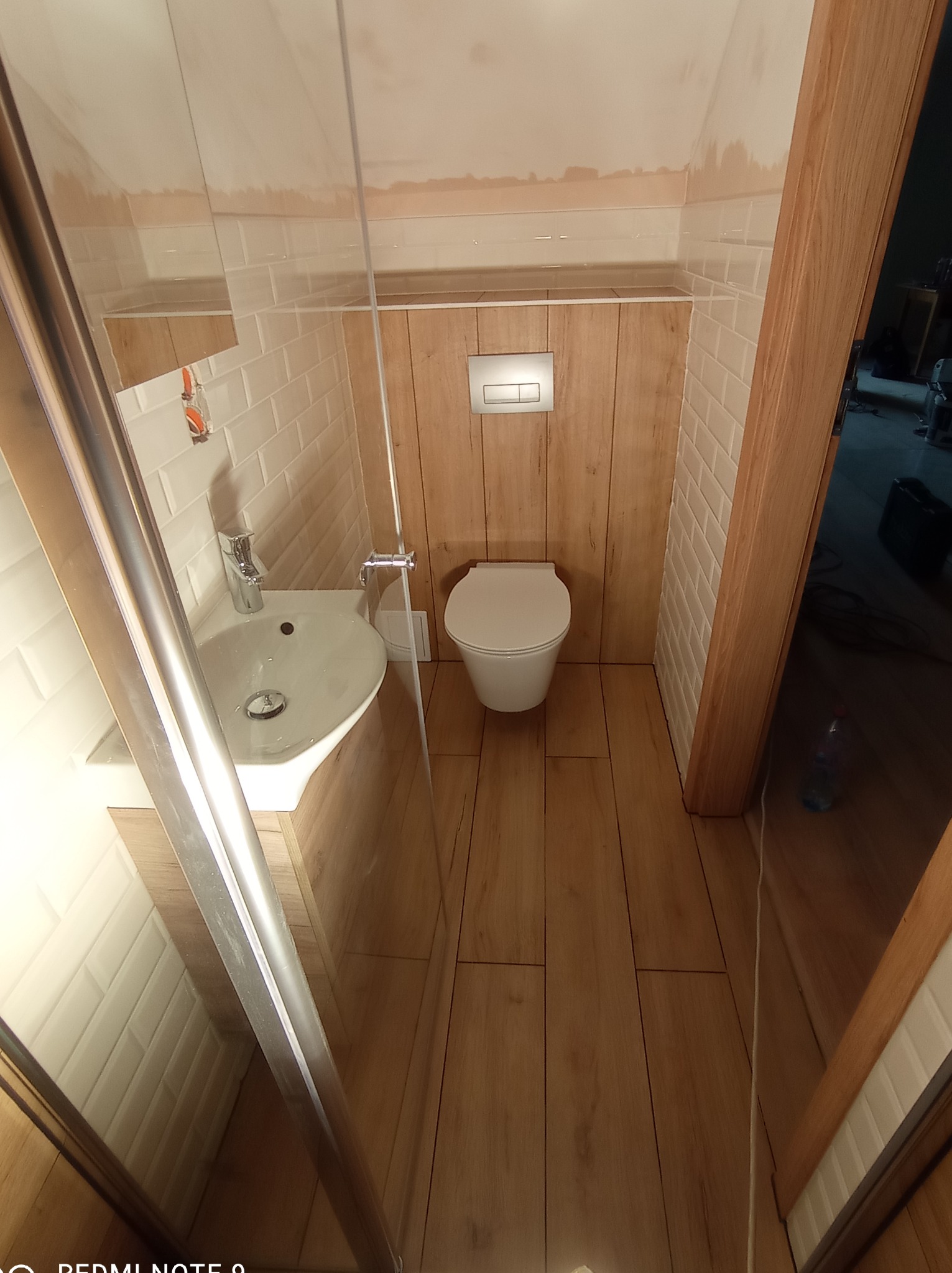 Wąska łazienka po remoncie z kabiną prysznicową, umywalką, toaletą i drewnianą boazerią do połowy ściany, wykończona białymi, prostokątnymi płytkami.