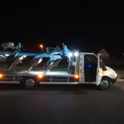 Nocny widok białej lawety z otwartymi drzwiami kabiny, przewożącej niebieski pług rolniczy z dodatkowym kołem na g&oacute;rze, oświetlonej pomarańczowymi światłami pozycyjnymi.