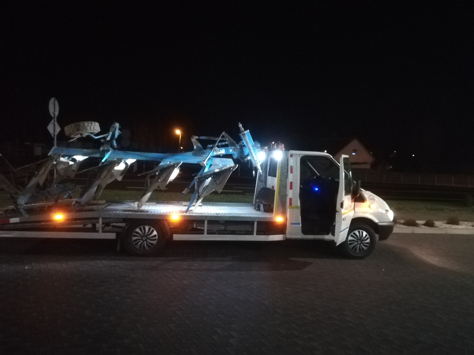 Nocny widok białej lawety z otwartymi drzwiami kabiny, przewożącej niebieski pług rolniczy z dodatkowym kołem na górze, oświetlonej pomarańczowymi światłami pozycyjnymi.