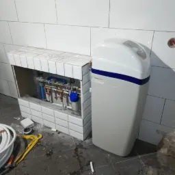 Filtr do wody obok zabudowanej instalacji hydraulicznej w pomieszczeniu z białymi płytkami, widoczne narzędzia i rury.