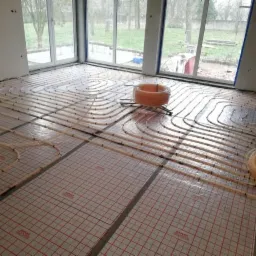 Rozłożone rury systemu grzewczego na podłodze z folią rastrową w trakcie instalacji; zwój rur w tle.