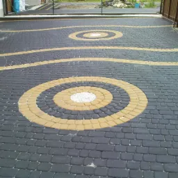 Czarno-żółta kostka brukowa ułożona we wzór fal i okręgów, z centralnym okręgiem wypełnionym białym żwirem.