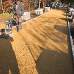 Pracownik ubija piaskowe podłoże pod przyszłą nawierzchnię chodnika przy użyciu zagęszczarki w słoneczny dzień.