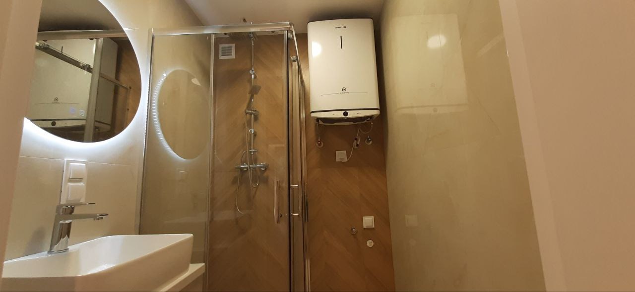 Nowoczesna łazienka po remoncie: kabina prysznicowa z transparentnego szkła, umywalka z baterią, podświetlane lustra, bojler na ścianie z imitacją drewna.