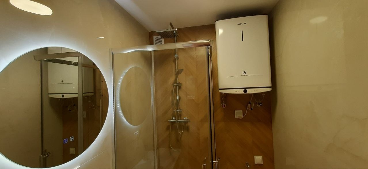 Wykończona łazienka z kabiną prysznicową, lustrem z podświetleniem LED i bojlerem na ścianie z panelami imitującymi drewno. Estetyczne wykończenie.
