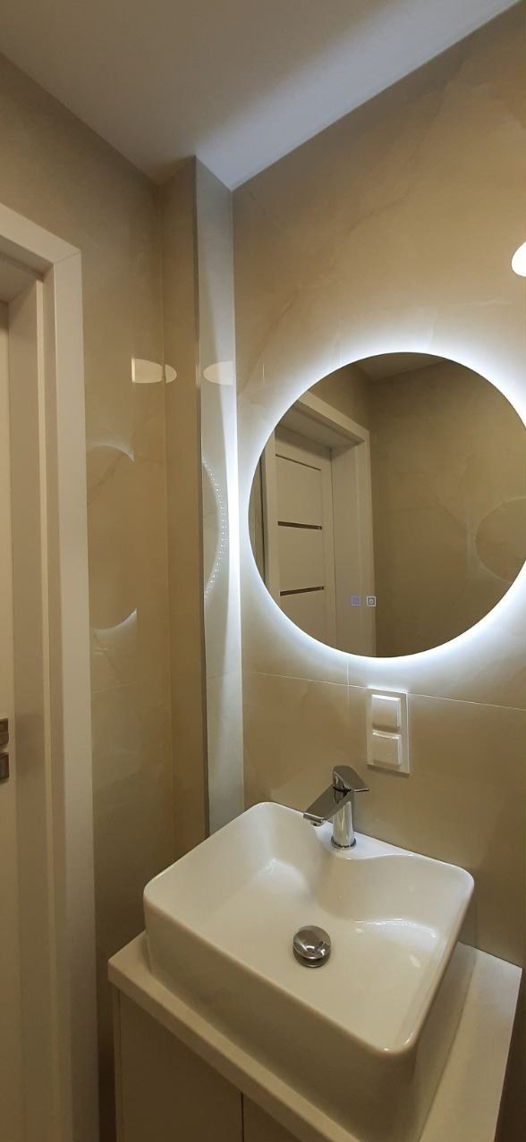 Beżowa łazienka po remoncie: umywalka nablatowa, okrągłe lustro z ledowym podświetleniem, kremowe płytki na ścianach i białe drzwi w tle.