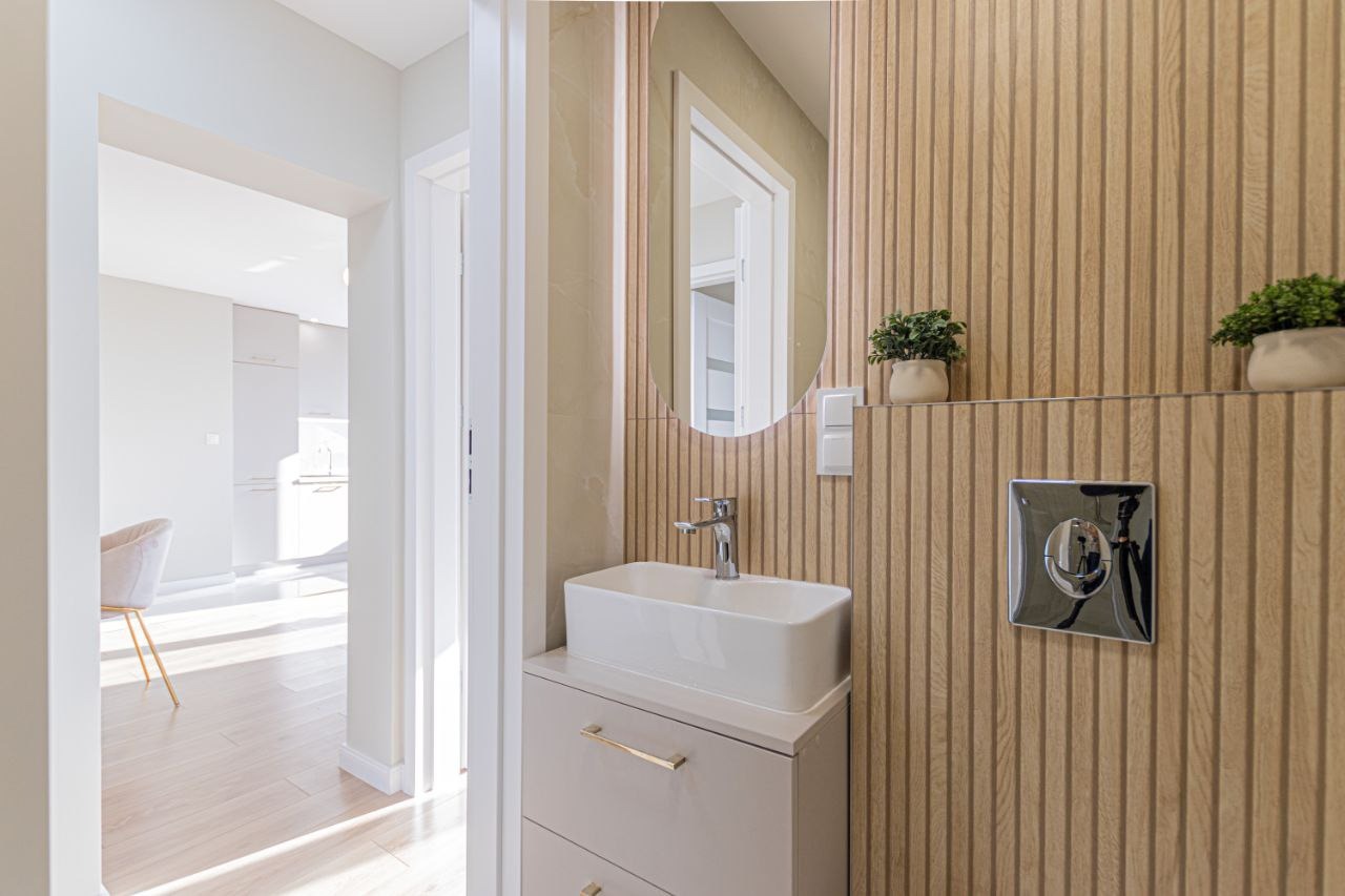 Nowoczesna łazienka z umywalką nablatową, okrągłym lustrem i drewnianą okładziną ścienną z roślinami. Widok na jasne wnętrze mieszkania.