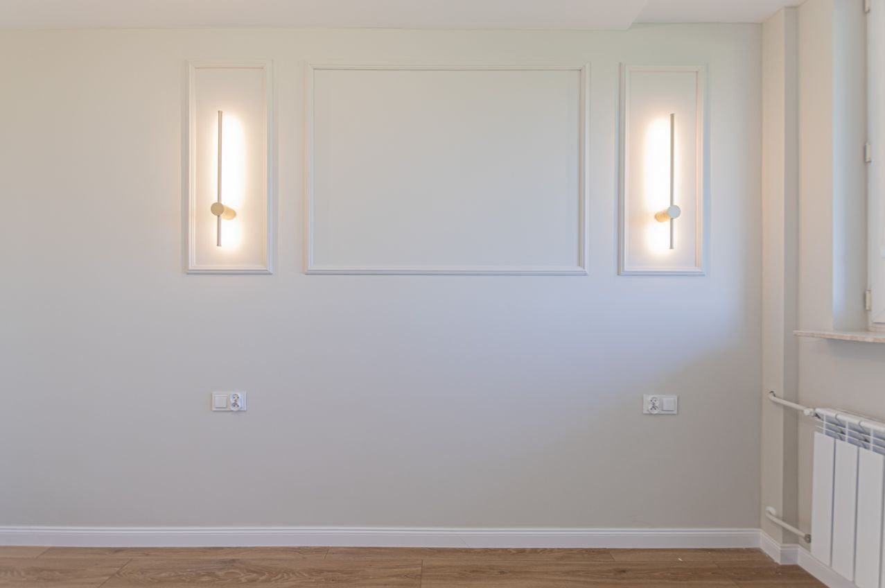 Ściana po szpachlowaniu i malowaniu z ozdobnymi ramami i nowoczesnymi kinkietami. Widoczne gniazdka elektryczne i grzejnik. Podłoga wykończona panelami.