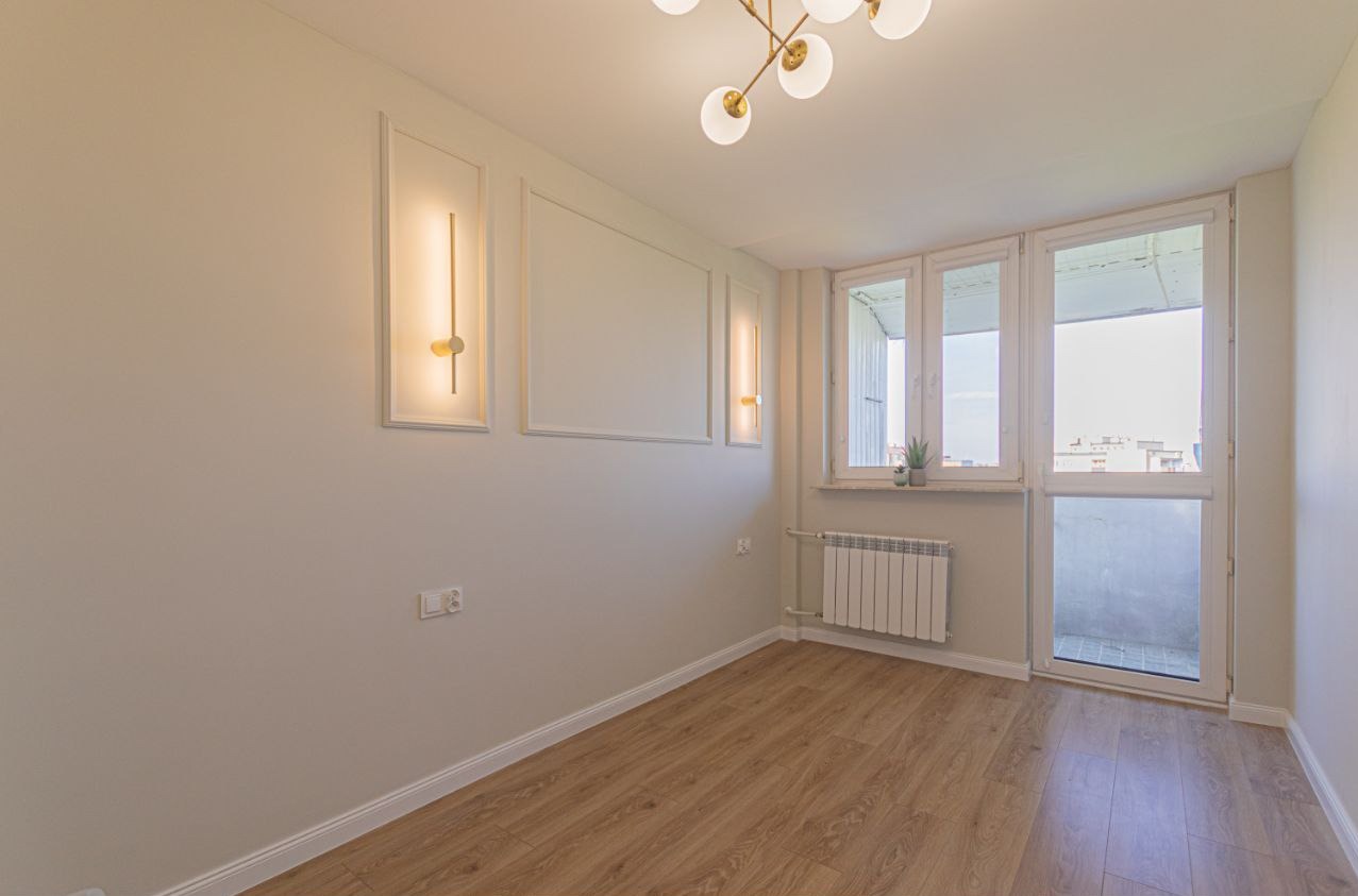 Jasne wnętrze po remoncie: drewnopodobna podłoga, białe ściany z dekoracyjnymi listwami i oświetleniem, okno balkonowe z widokiem na miasto, nowoczesna lampa sufitowa.