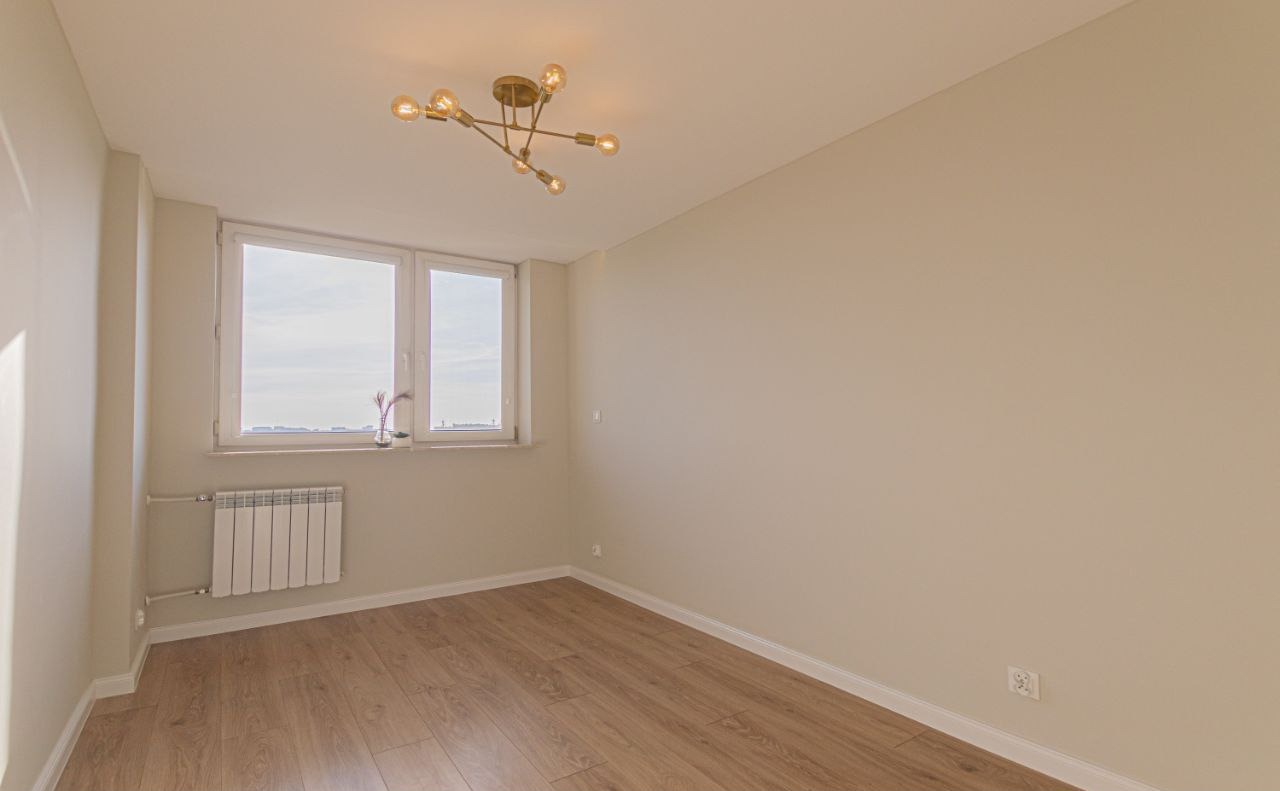 Jasne, puste wnętrze po remoncie: panele podłogowe, ściany w odcieniach beżu, okno z grzejnikiem i nowoczesna lampa sufitowa.