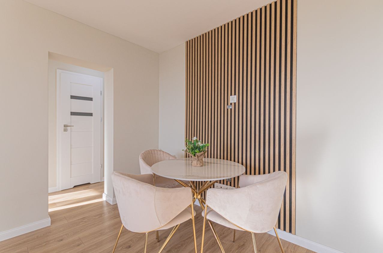 Jasne wnętrze jadalni z okrągłym stołem na złotych nogach, welurowe krzesła i dekoracyjna ściana z drewnianych lameli. W tle widoczne drzwi.