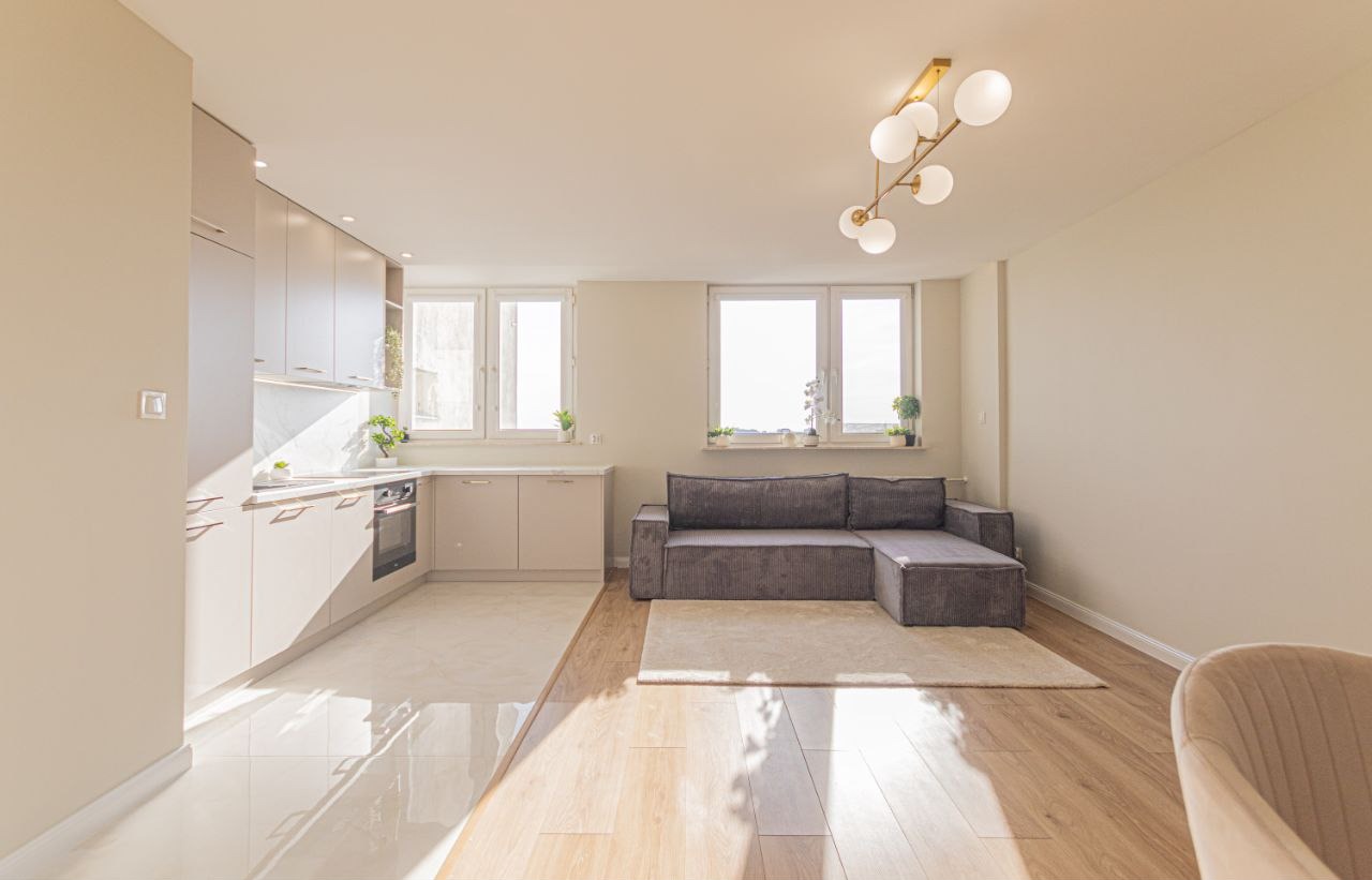 Jasne wnętrze mieszkania po remoncie: kuchnia z beżowymi szafkami i salon z szarą sofą. Parkiet i płytki podłogowe oddzielają strefy. Nowoczesna lampa sufitowa.