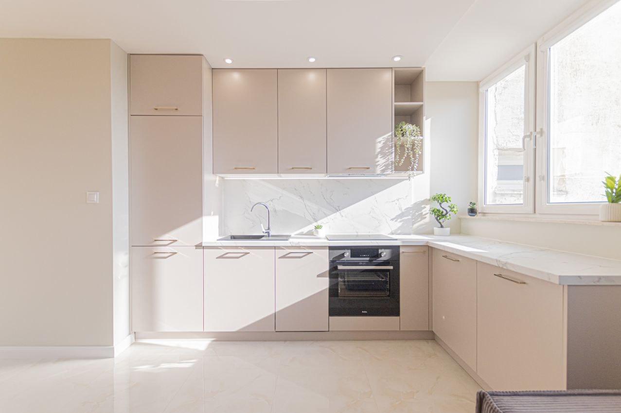 Jasna kuchnia z beżowymi szafkami, marmurowym blatem i oświetleniem punktowym. Minimalistyczny design z roślinami doniczkowymi i dużymi oknami.