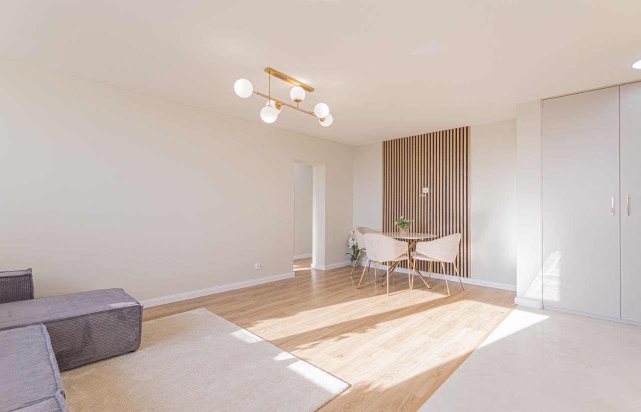 Jasne wnętrze po remoncie: parkiet, drewniana ściana, okrągły stół z krzesłami i nowoczesna lampa sufitowa. Minimalistyczny design.