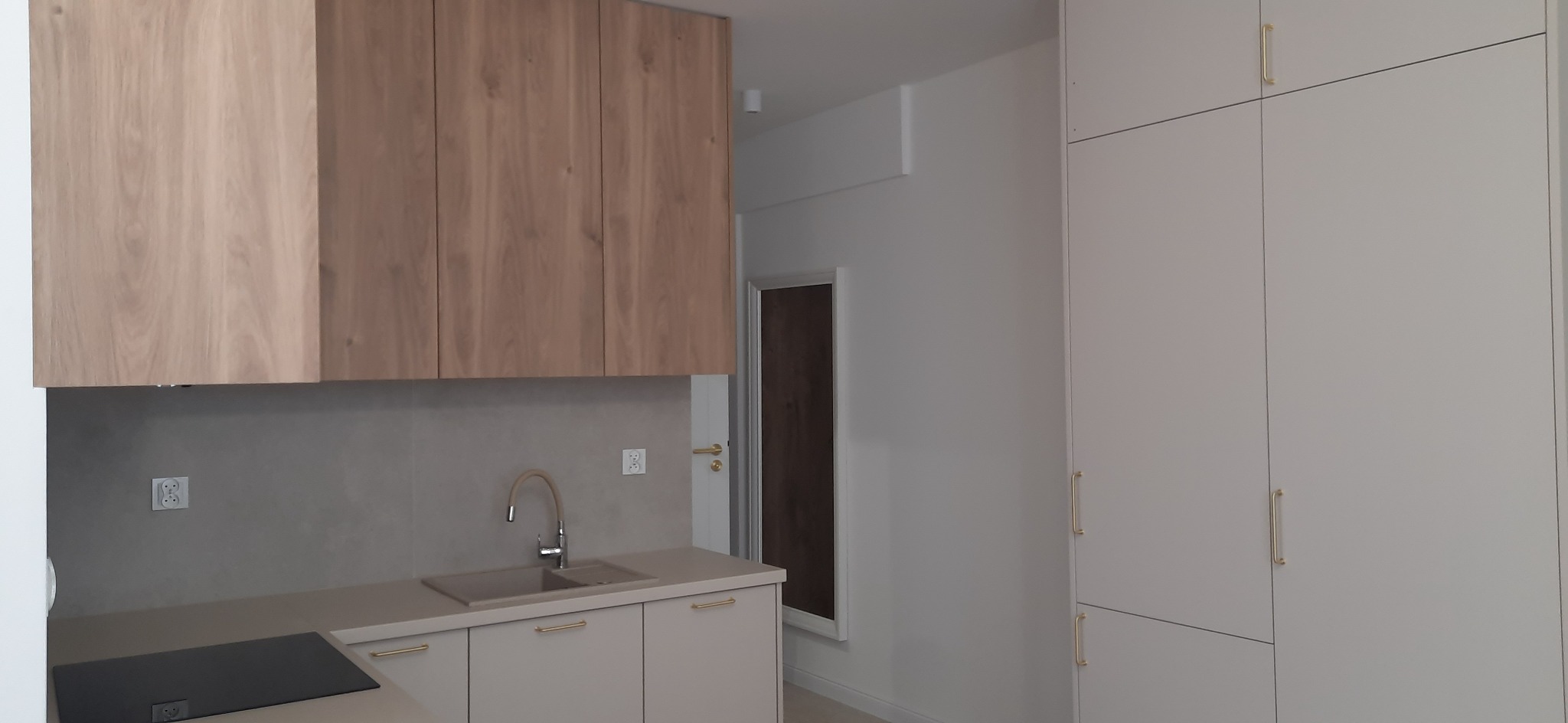 Jasna, nowoczesna kuchnia z drewnianymi szafkami górnymi i białymi dolnymi, minimalistyczny design, złote uchwyty, płytki ścienne w odcieniach szarości.