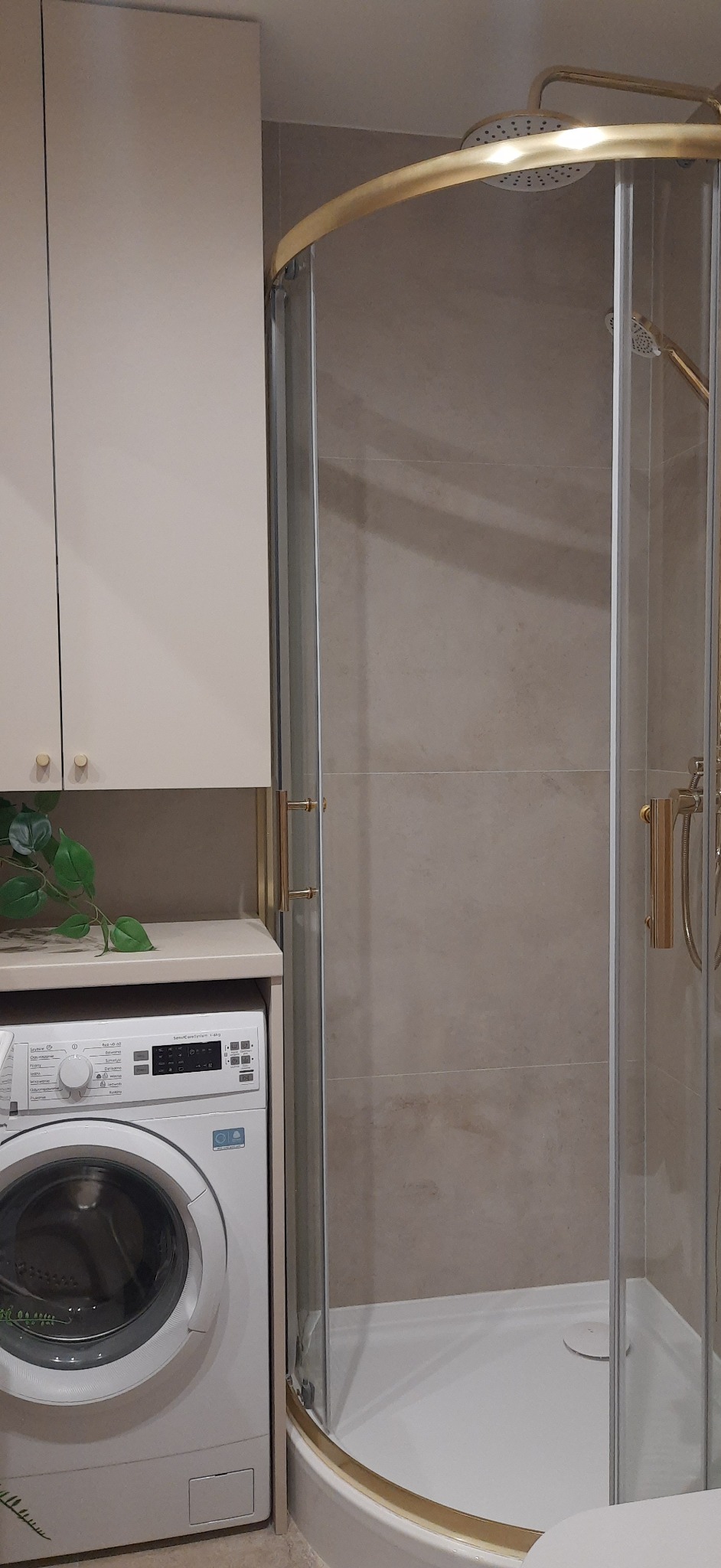 Wykończona łazienka z kabiną prysznicową z złotymi elementami i pralką w zabudowie. Beżowe płytki na ścianach i szafka nad pralką. Nowoczesny design.