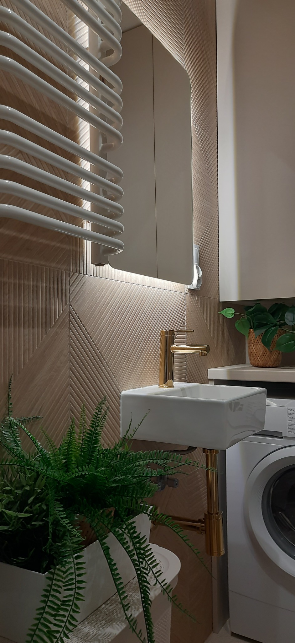 Nowoczesna łazienka w Lublinie: beżowe płytki z deseniem, biała umywalka na złotym stelażu, złota bateria, grzejnik drabinkowy i rośliny doniczkowe tworzą spójną całość.