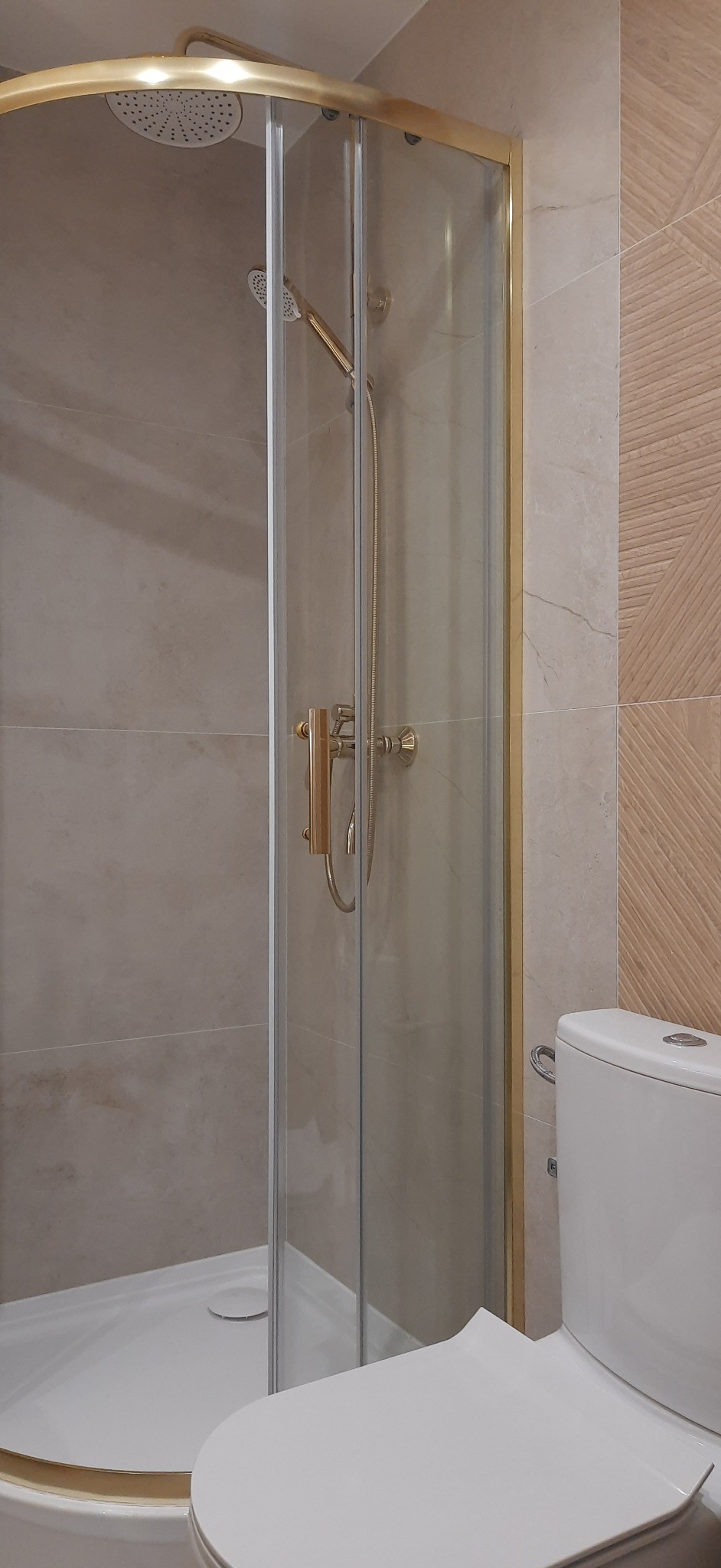 Kabina prysznicowa z brodzikiem i złotymi detalami, widoczna obok toaleta. Płytki ścienne w beżach i brązach tworzą nowoczesną aranżację łazienki.