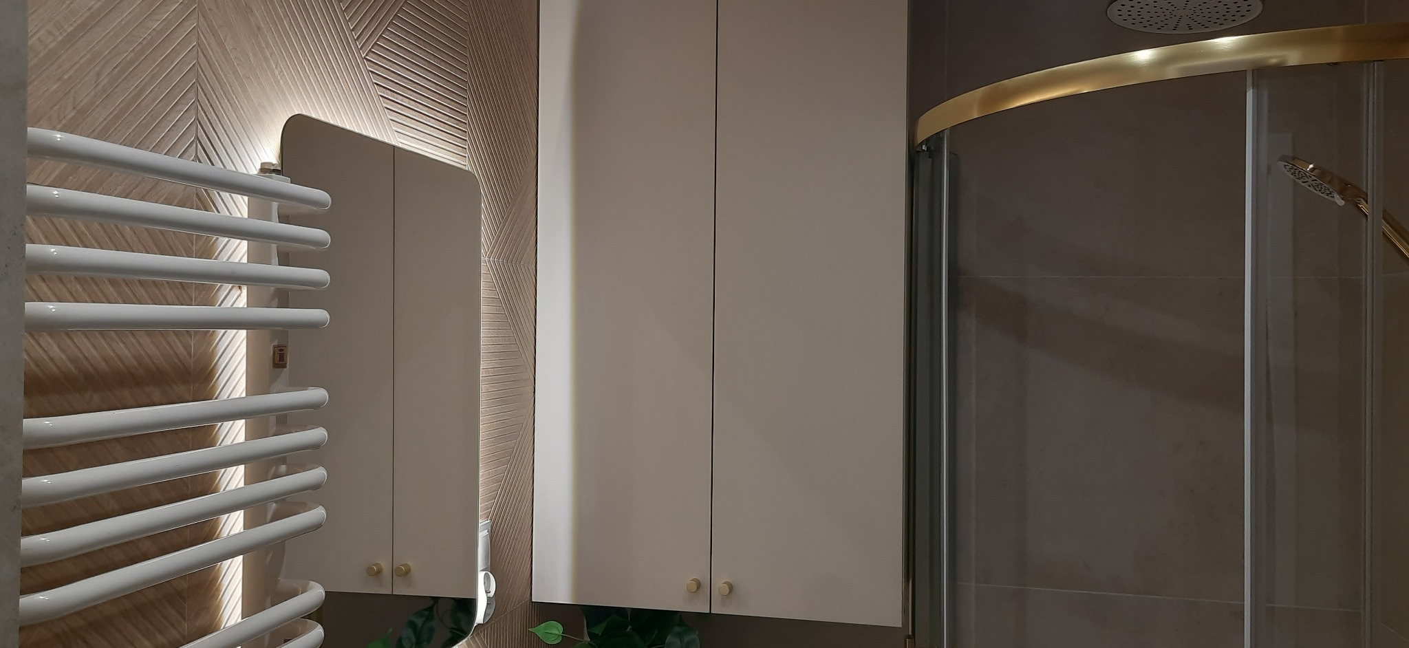 Nowoczesna łazienka z grzejnikiem drabinkowym, szafkami w kolorze ecru i kabiną prysznicową z elementami w kolorze złotym. Na ścianie dekoracyjne panele.