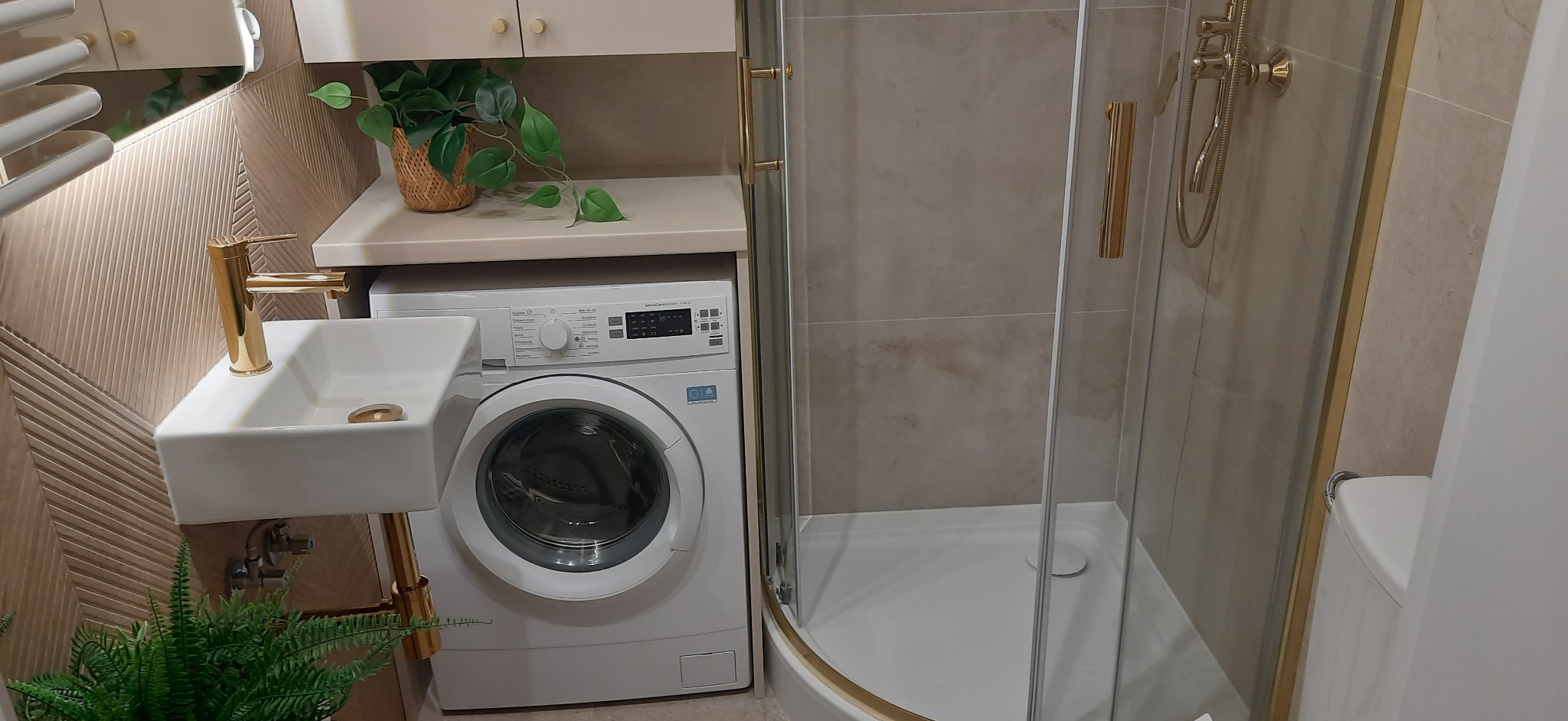 Nowoczesna łazienka z kabiną prysznicową, umywalką na blacie i pralką pod blatem. Złote akcenty armatury i detali. Neutralna kolorystyka płytek i mebli.