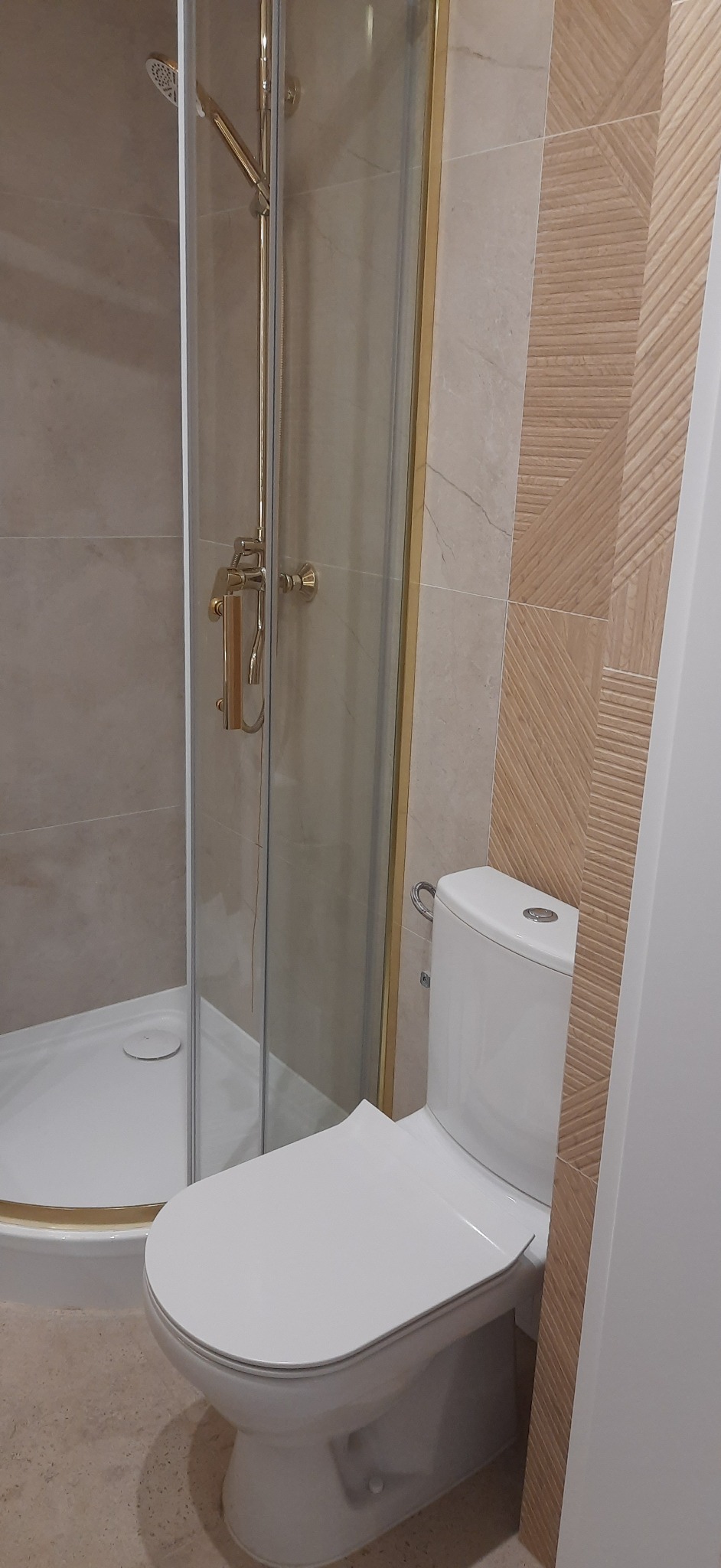 Wykończona łazienka z kabiną prysznicową w złotej ramie i toaletą. Na ścianie dekoracyjne płytki w odcieniach beżu i brązu. Całość utrzymana w jasnej kolorystyce.