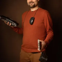 Uśmiechnięty mężczyzna prezentuje kartę graficzną w jednej ręce i kartę dźwiękową z wentylatorem w drugiej, z myszką komputerową zawieszoną na szyi.