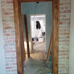 Drewniana ościeżnica drzwiowa w trakcie montażu, widoczna surowa cegła na ścianie i narzędzia w tle, wnętrze w trakcie remontu.