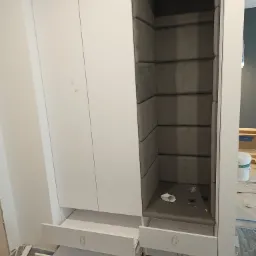 Biała szafa wnękowa z otwartymi szufladami i tapicerowanym wnętrzem przypominającym siedzisko, widoczne drobne zabrudzenia na podłodze i ślady prac wykończeniowych.