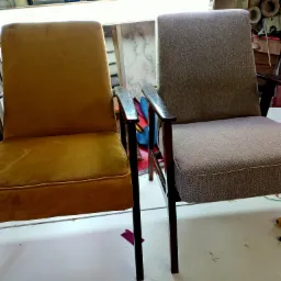 Dwa odnowione fotele z drewnianymi podłokietnikami, jeden w odcieniu musztardowym, drugi w szarym, stoją obok siebie na białym stole w warsztacie z narzędziami w tle.