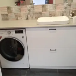 Nowoczesna łazienka z pralką LG obok białej szafki umywalkowej z ciemnymi uchwytami, lustro odbija osobę robiącą zdjęcie.