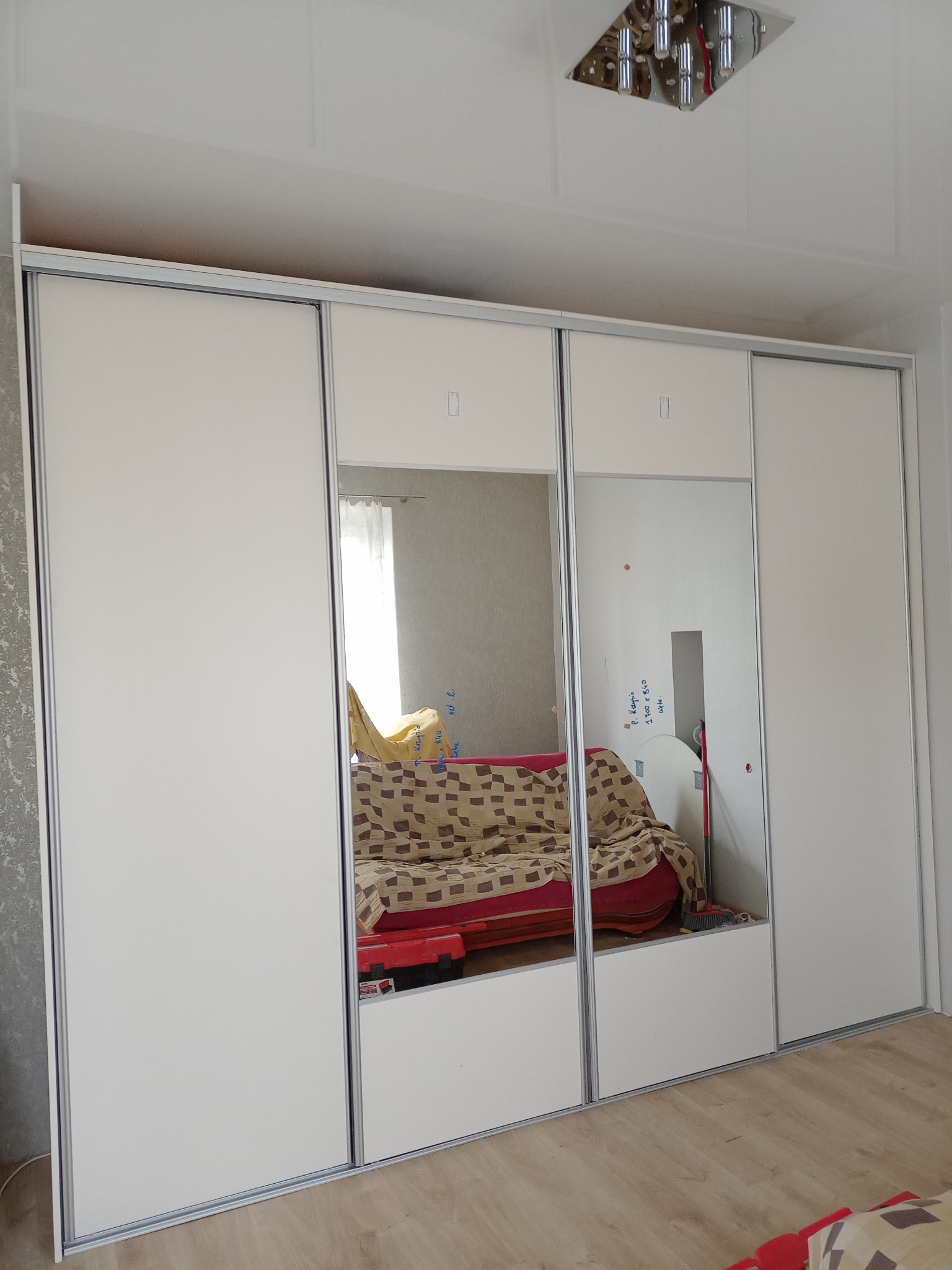 Biała szafa przesuwna z lustrami, odbijająca fragment pokoju z sofą i napisanymi na ścianie wymiarami.