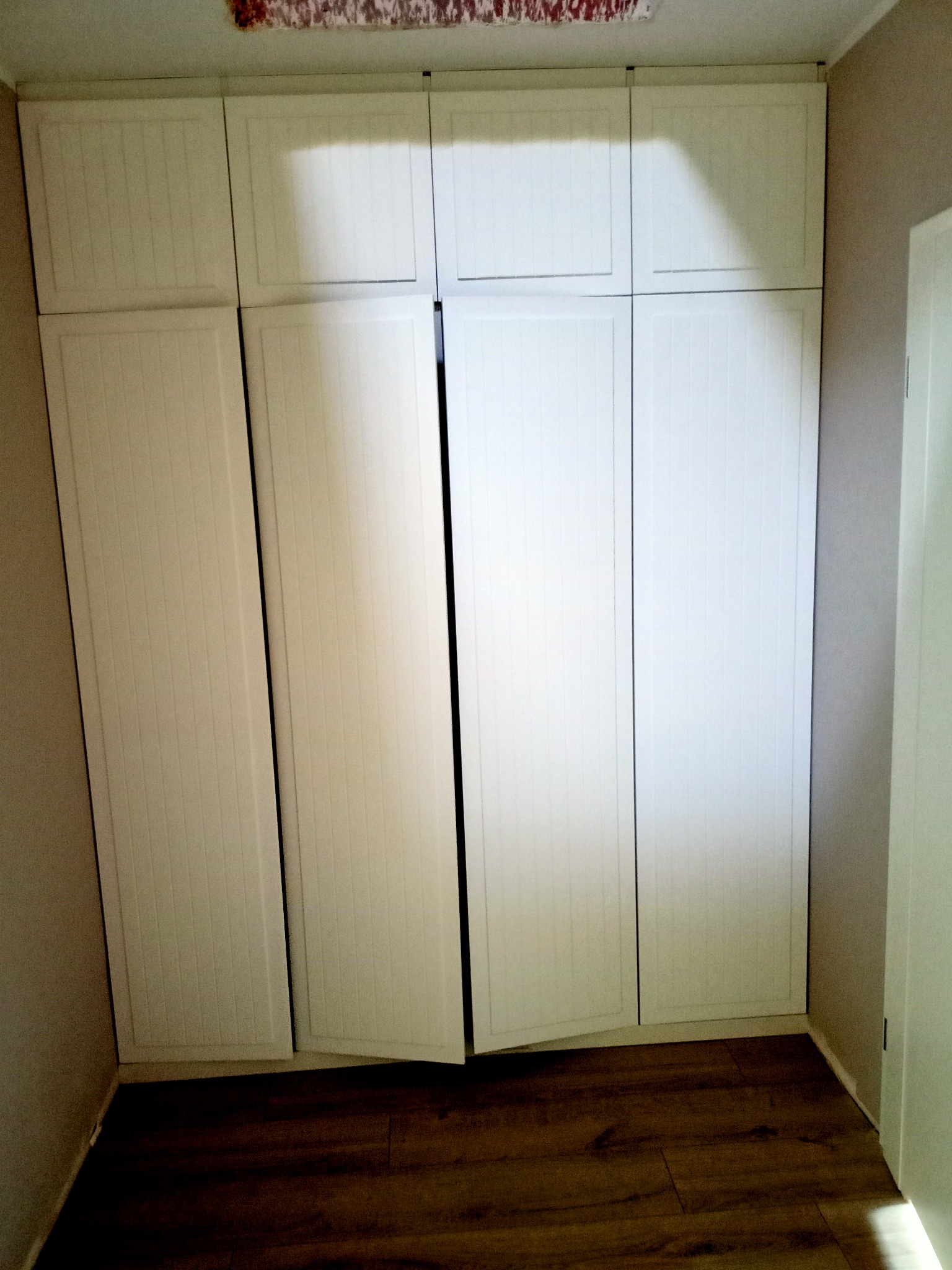 Biała szafa wnękowa z frontami z pionowymi żłobieniami, jedna para drzwi lekko otwarta, widoczna drewniana podłoga i fragment ściany.