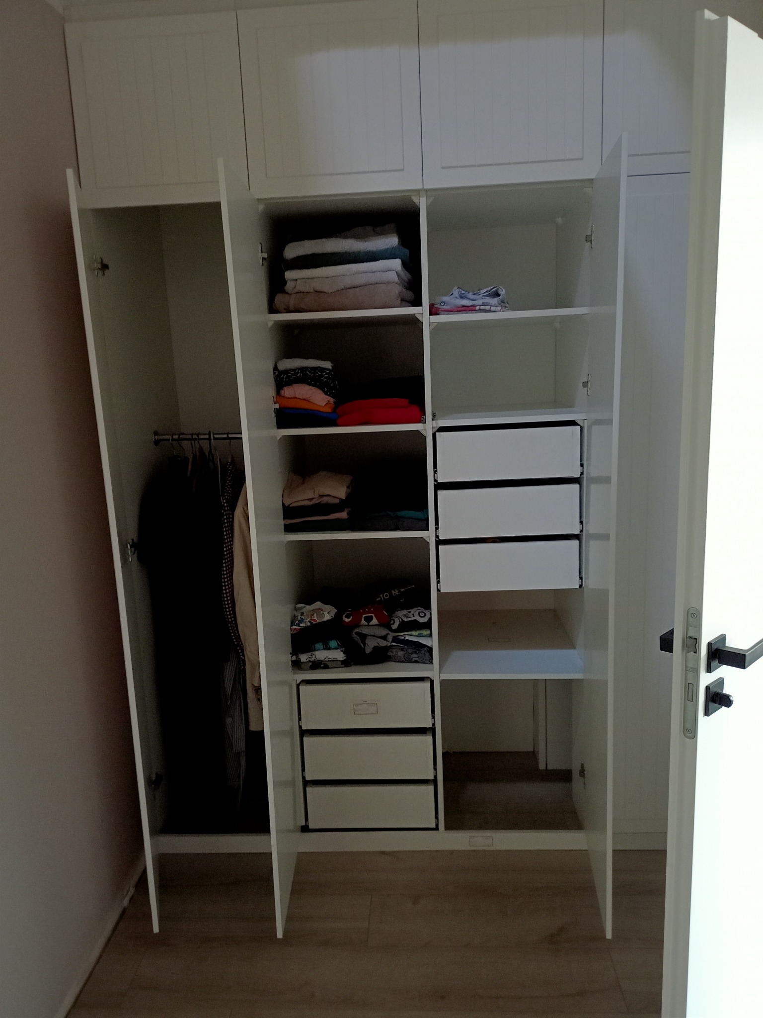 Wnętrze białej, trzydrzwiowej szafy z półkami, szufladami i drążkiem na ubrania, wypełnione ubraniami i tekstyliami.