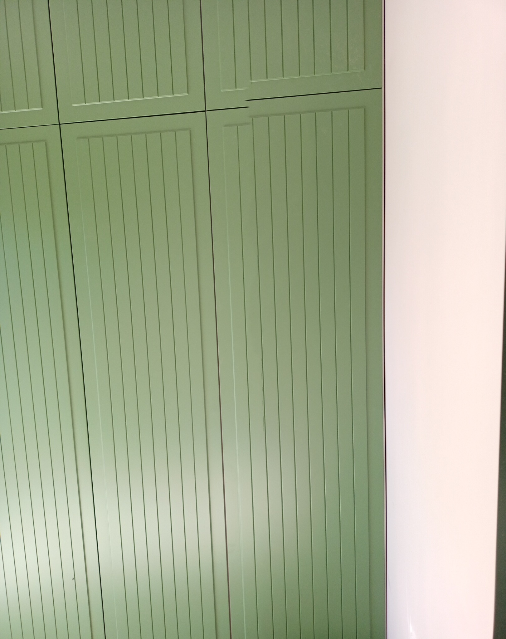 Fragment szafy wnękowej z zielonymi, frezowanymi frontami, przylegającej do białej ściany.