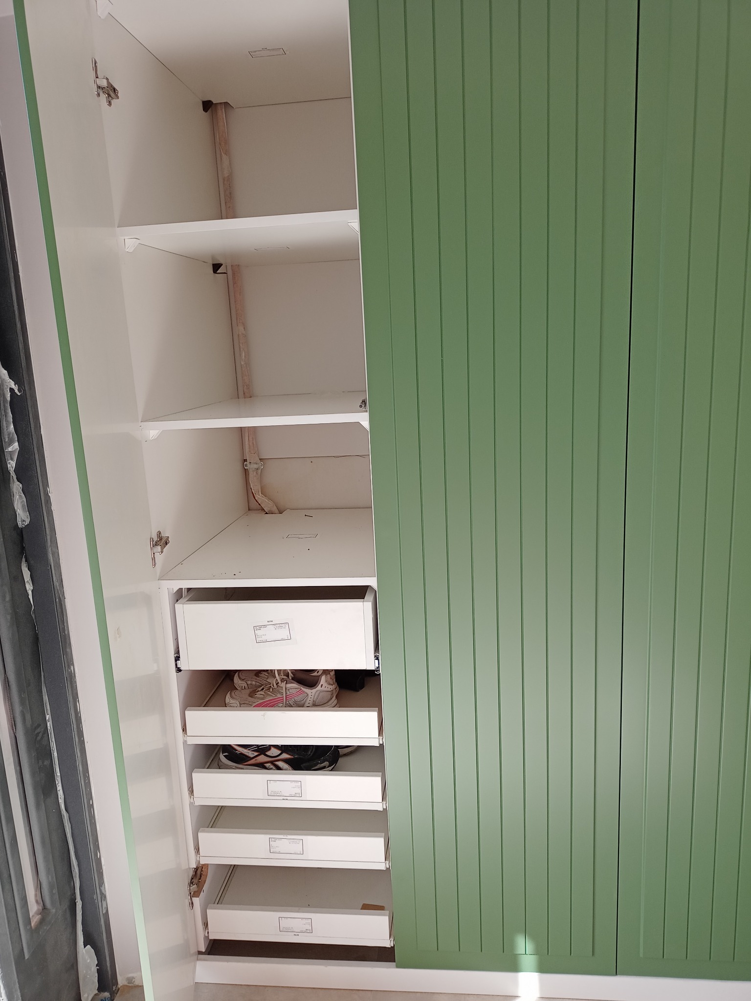 Otwarta szafa z białymi półkami i szufladami, częściowo wypełniona obuwiem, z zielonymi, pionowo żłobkowanymi drzwiami.