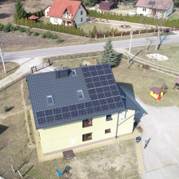 ARCHIGON Sp. z o.o. - Widok z lotu ptaka na dom z częściowo zamontowanymi panelami słonecznymi na dachu, obok drugi zestaw paneli na ziemi, w tle zabudowa jednorodzinna i plac zabaw.