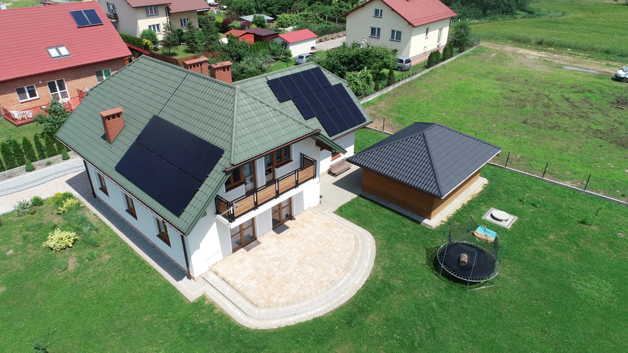 Widok z drona na dom z zielonym dachem i panelami fotowoltaicznymi, obok drewniana altana z szarym dachem i trampolina.