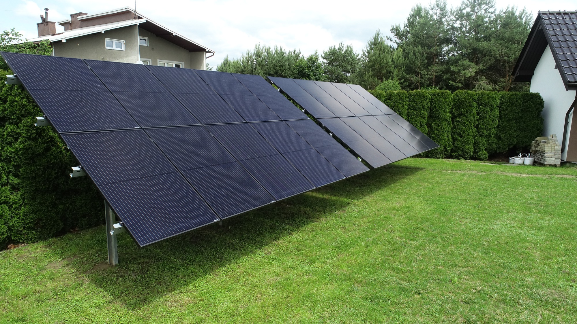 Dwa rzędy paneli słonecznych ustawione na trawniku przed domem jednorodzinnym z zielonym żywopłotem.