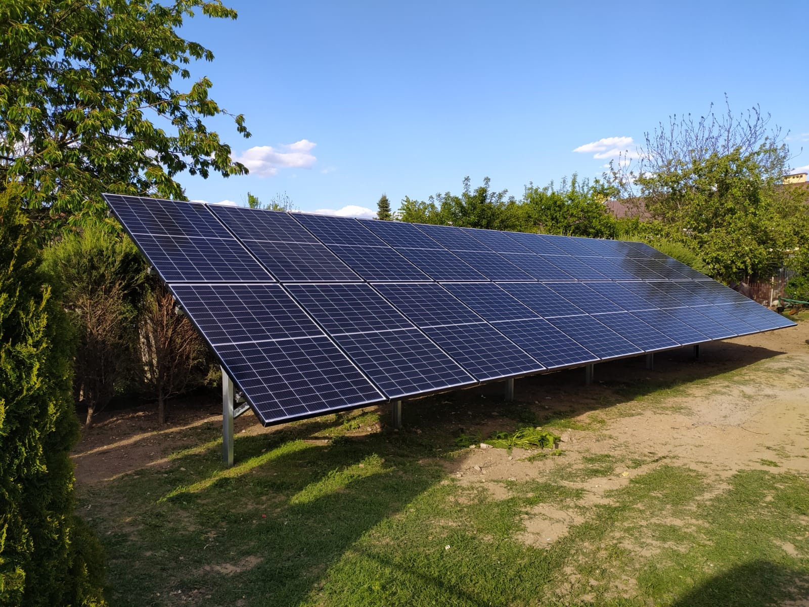 Instalacja paneli słonecznych na metalowej konstrukcji, widok z boku, w tle zieleń drzew i błękitne niebo.