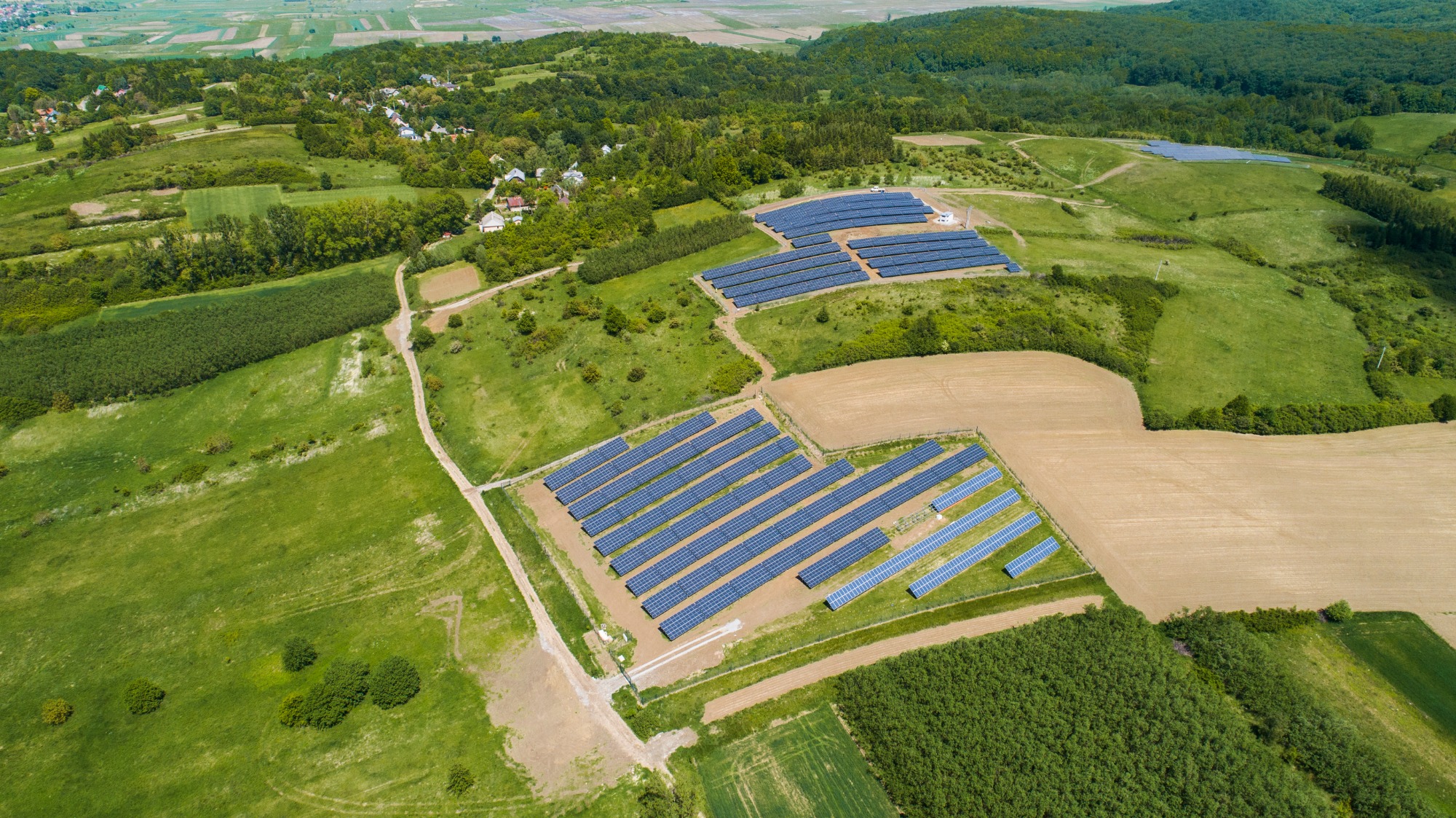 Widok z lotu ptaka na farmę paneli słonecznych na wzgórzu, otoczoną zielenią i lasami w oddali.