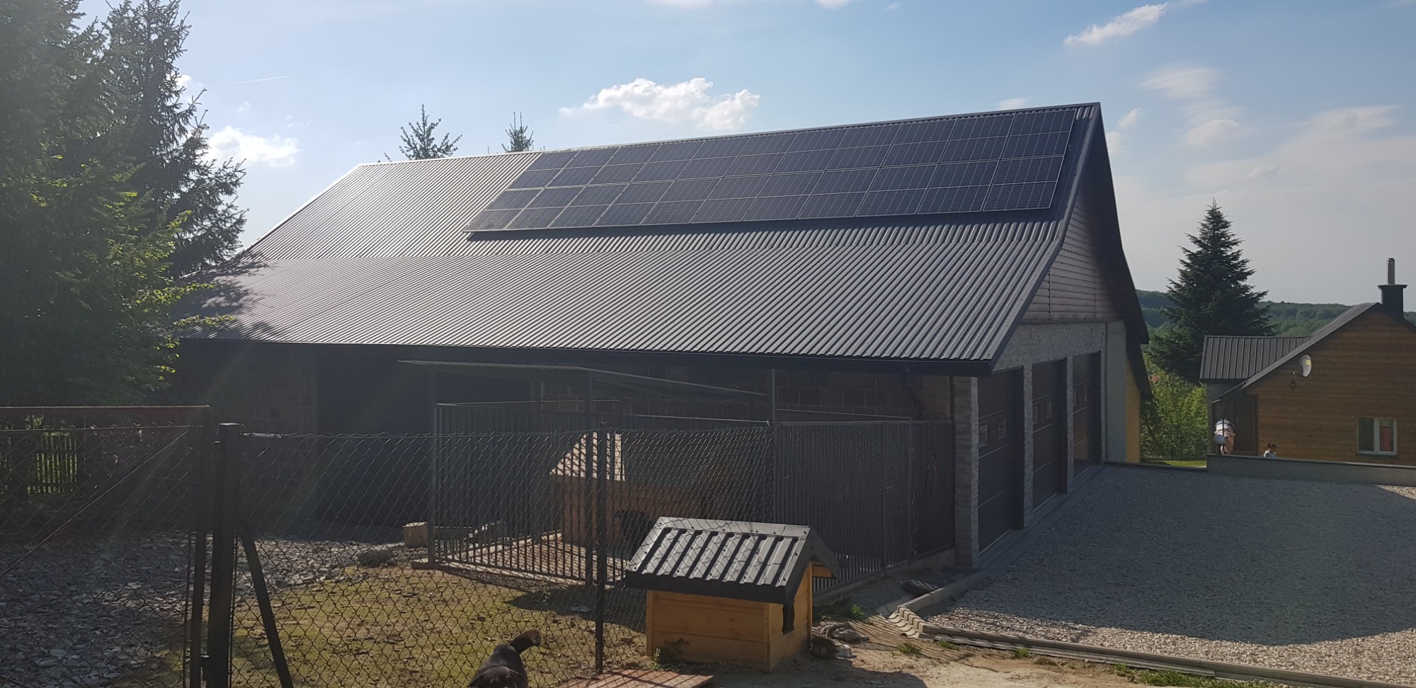 Dom z ciemnym, metalowym dachem, na którym zamontowane są panele słoneczne. Widoczny również kojec dla psa i buda.