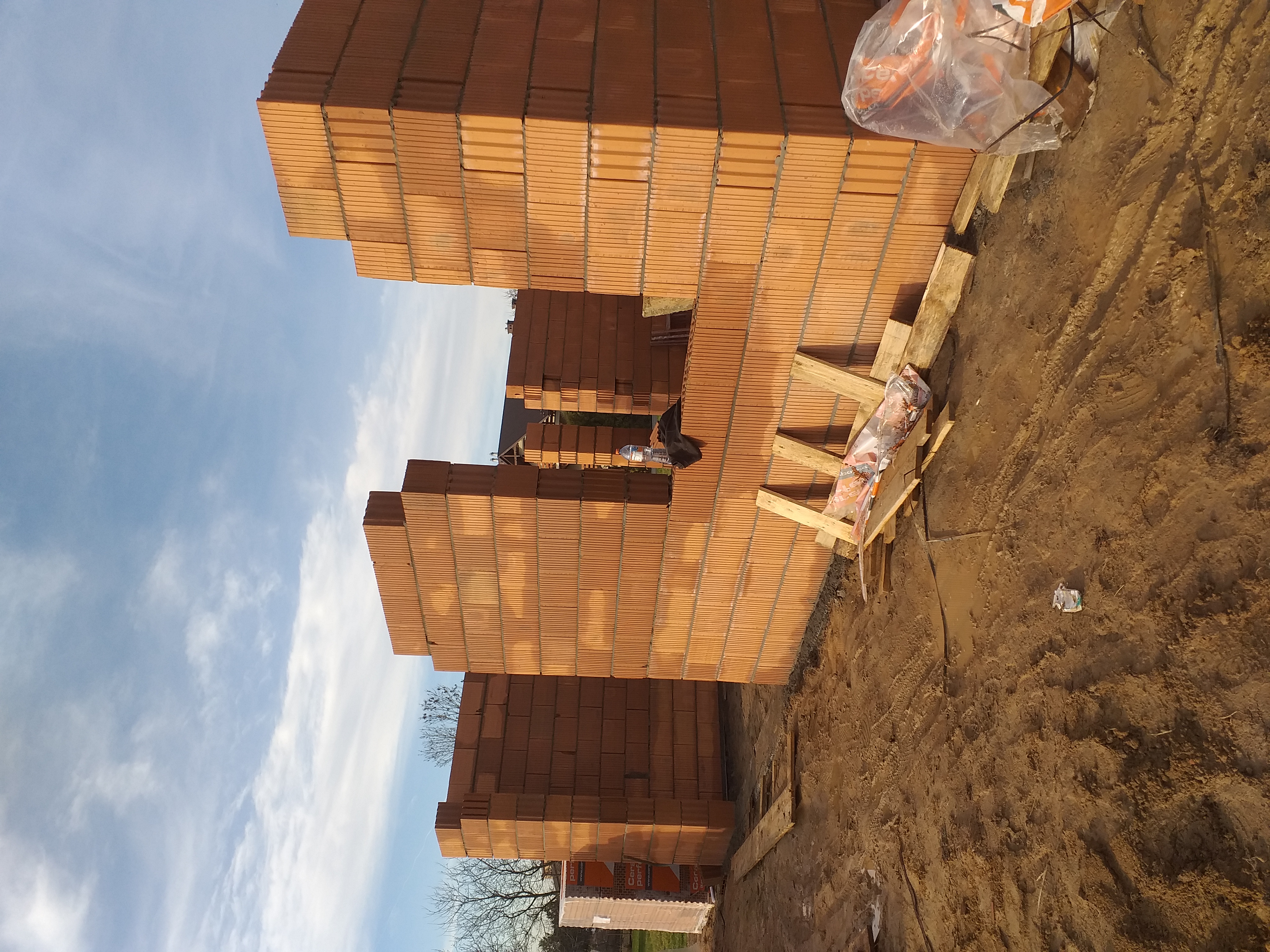 Wznoszone ściany fundamentowe z pustaków ceramicznych, widoczne zbrojenie i drewniane podpory, w tle błękitne niebo z chmurami.