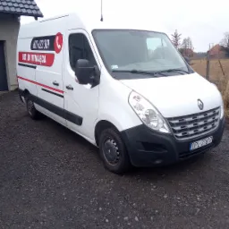 Biały Renault Master z numerem telefonu i napisem 'Bus do wynajęcia' na boku, zaparkowany na żwirowej nawierzchni przed budynkiem.